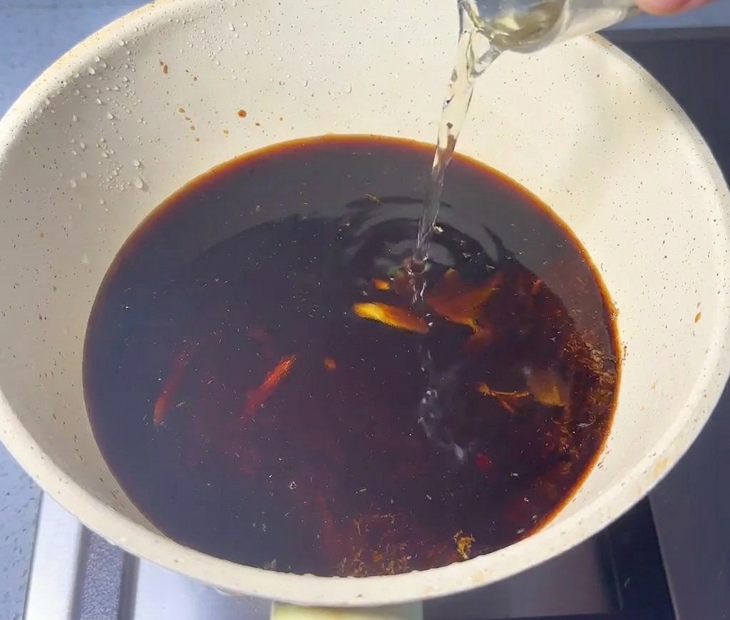 Helle Flüssigkeit wird aus einem Glas in einen Topf mit dunkler Sauce und Fischpulver gegossen.