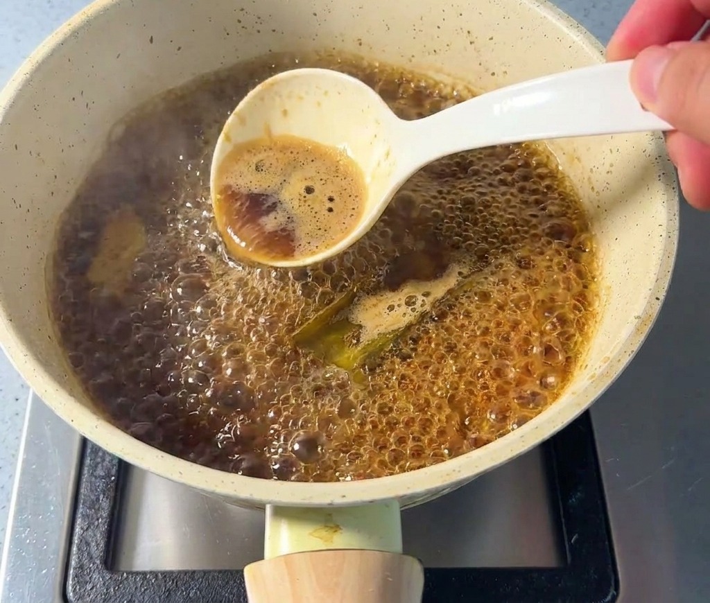 Eine Hand schöpft mit einem Löffel Schaum von der Oberfläche einer köchelnden dunklen Sauce ab.
