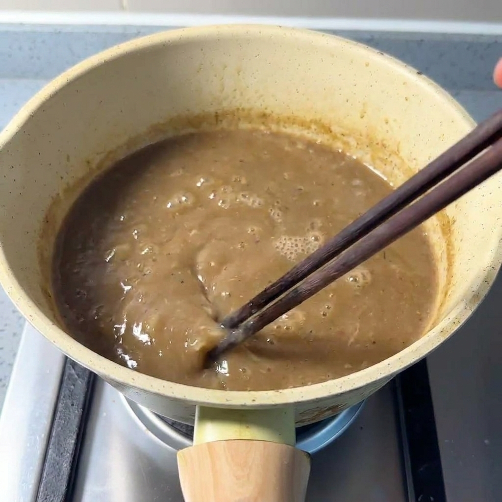 Eine dicke, bräunliche Brühe wird in einem Topf mit Stäbchen umgerührt.