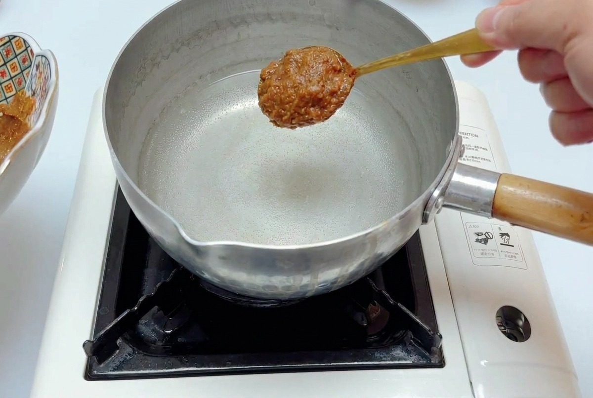 Ein Löffel mit dicker Miso-Würzpaste wird über einen Edelstahltopf mit klarem Wasser gehalten.