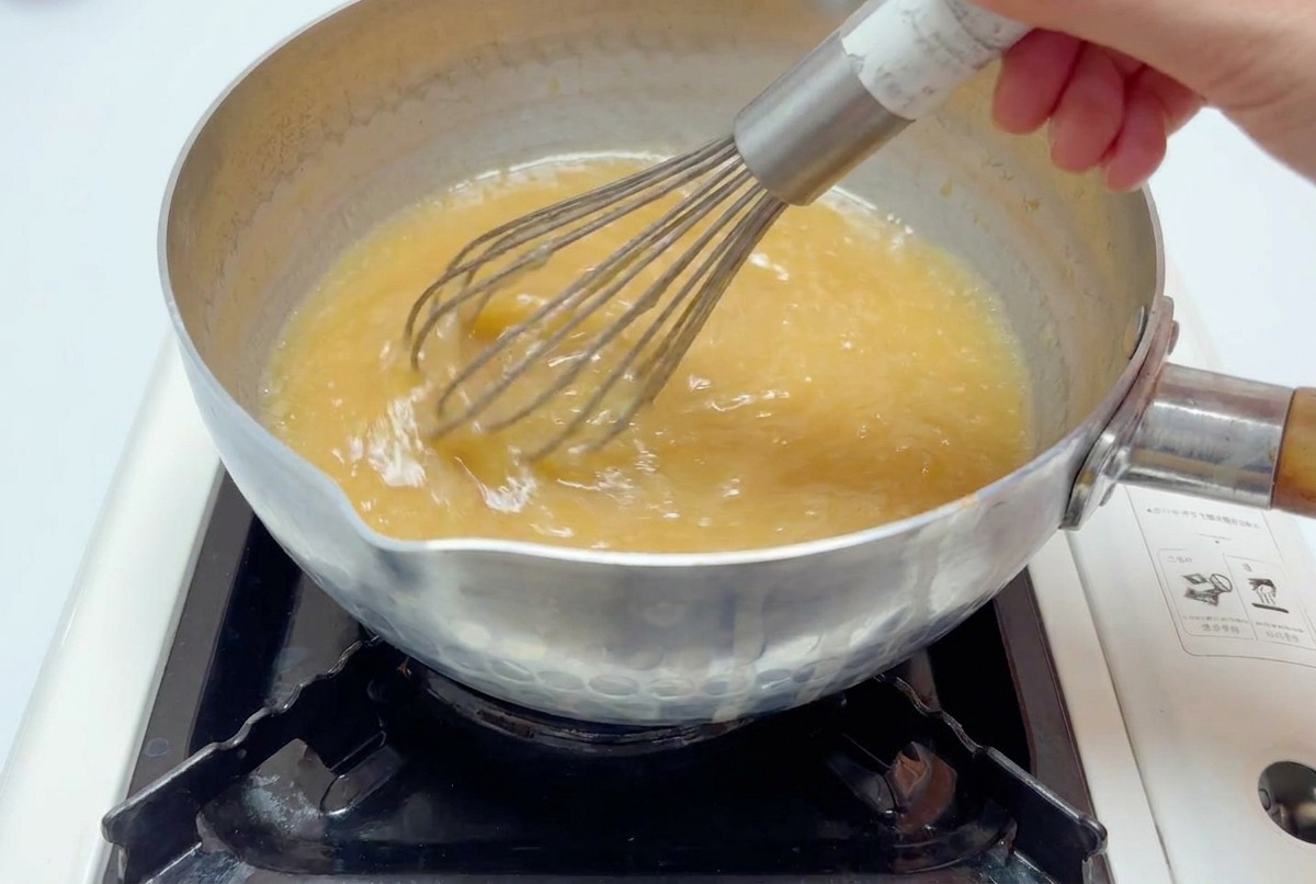 Ein Schneebesen rührt die hellbraune Miso-Brühe in einem Topf über einer Flamme glatt.