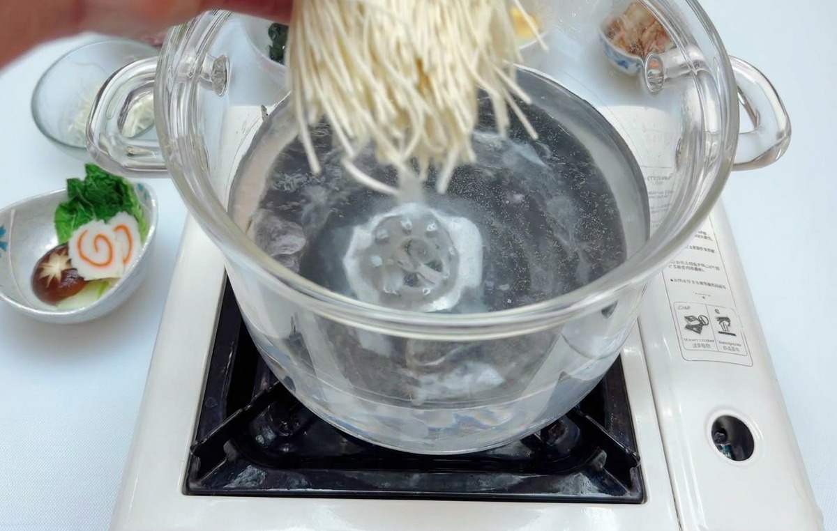 Eine Hand gibt ein Bündel weißer Ramennudeln in einen Topf mit sprudelnd kochendem Wasser.