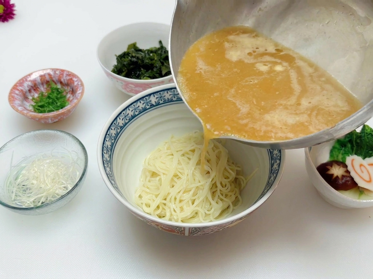 Heiße, goldbraune Miso-Suppe wird aus einem Edelstahltopf über die Ramennudeln gegossen.
