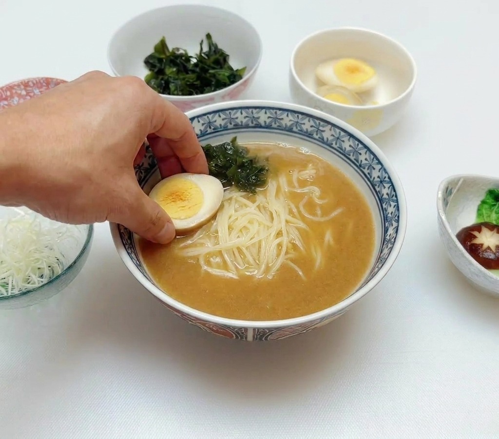 Eine Hand legt ein halbiertes weichgekochtes Ei auf die Ramen mit Brühe und Wakame.