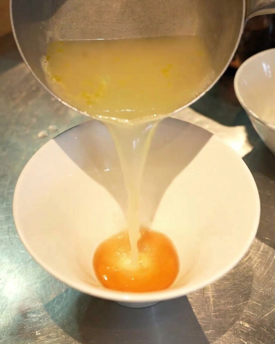 Ein Strahl klarer, goldener Hühnerbrühe wird aus einem Topf in eine Schüssel mit dunklerer Würzpaste gegossen.