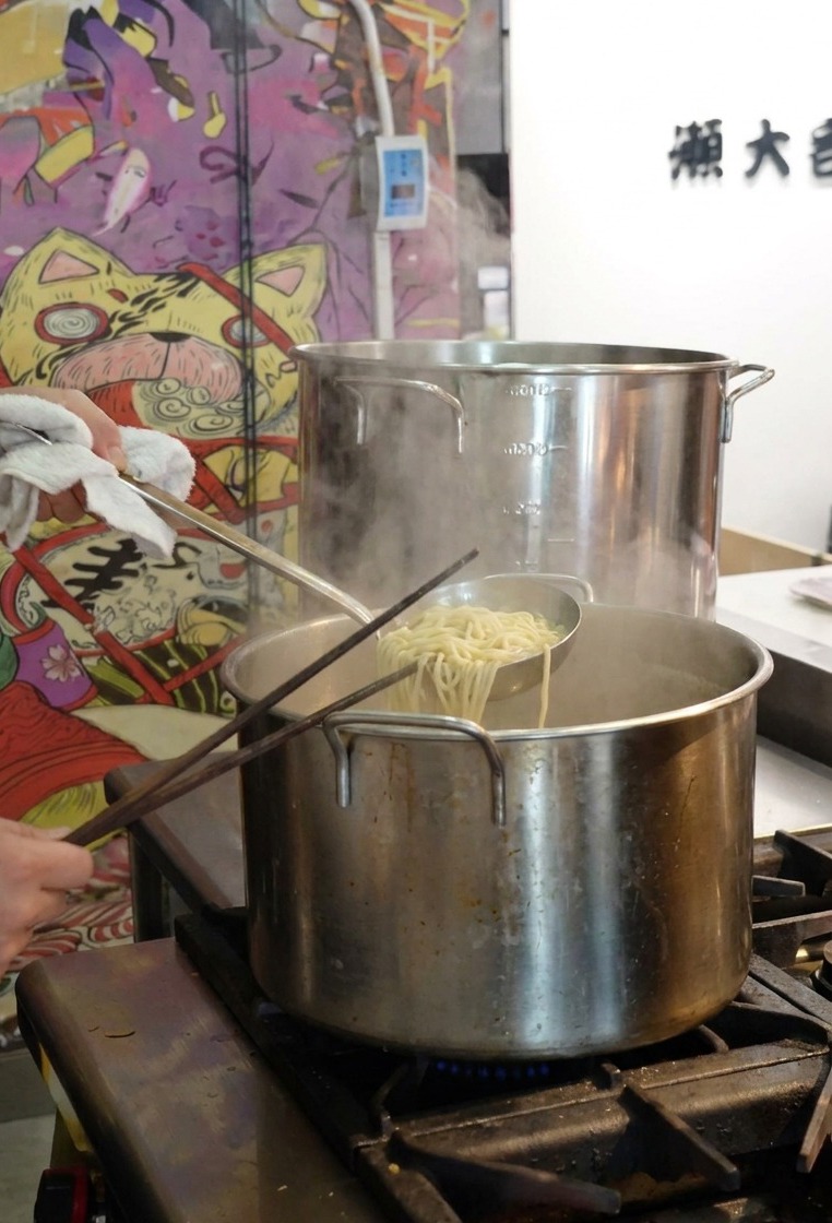 Eine Person hebt die gekochten Ramen-Nudeln mit einem Sieb aus dem Topf mit kochendem Wasser.