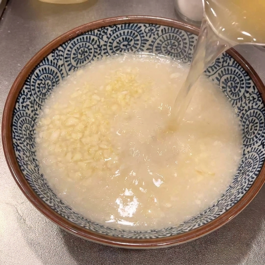 Heiße, klare Ramen-Brühe wird aus einem Topf in eine blau-weiße Keramikschale mit geriebenem Knoblauch gegossen.