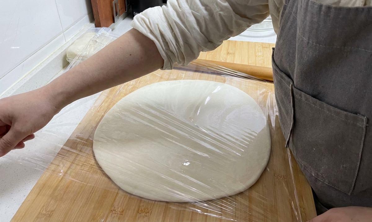 Eine Hand zieht Frischhaltefolie über eine flache Teigplatte auf einem Holzbrett.