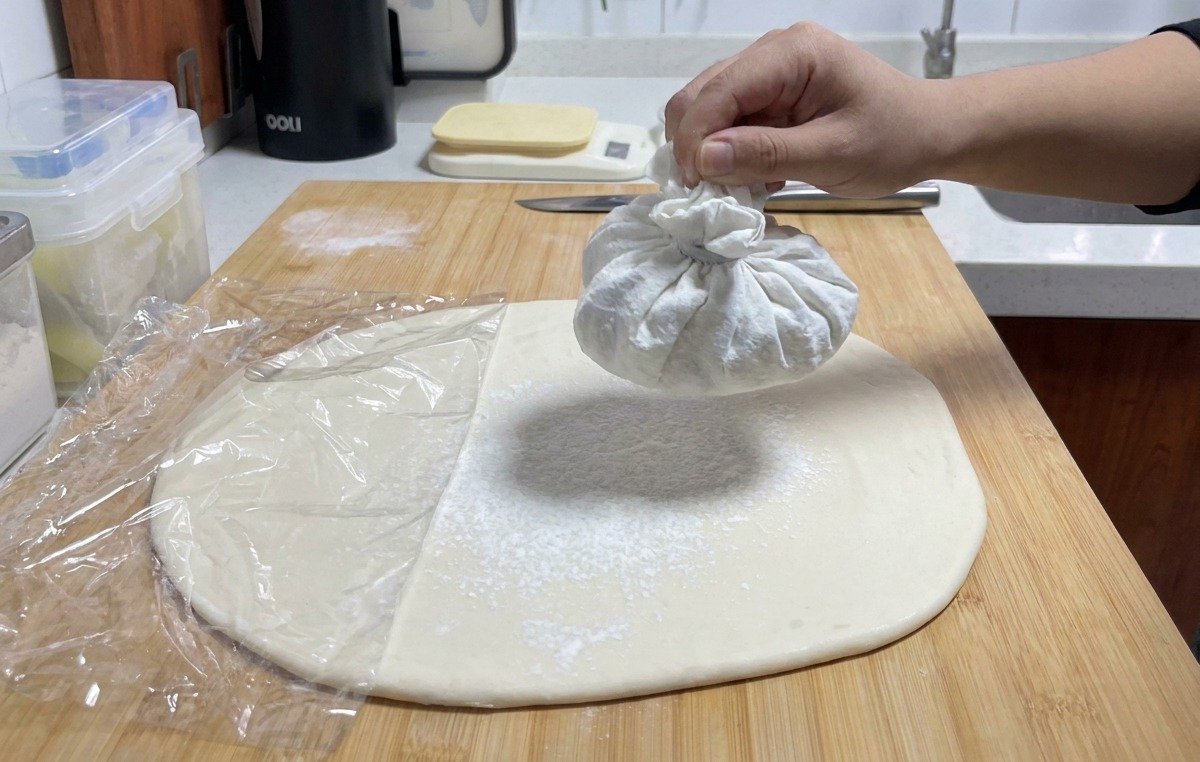 Eine Hand schüttelt ein Mehlsäckchen über der Teigplatte aus, um einen feinen Mehlnebel zu erzeugen.