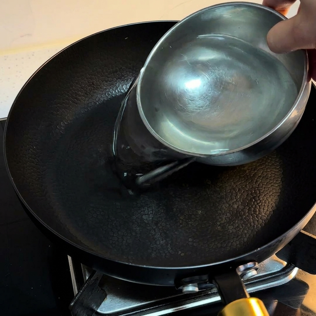 Wasser wird aus einer kleinen Edelstahlschüssel in eine schwarze Pfanne gegossen.