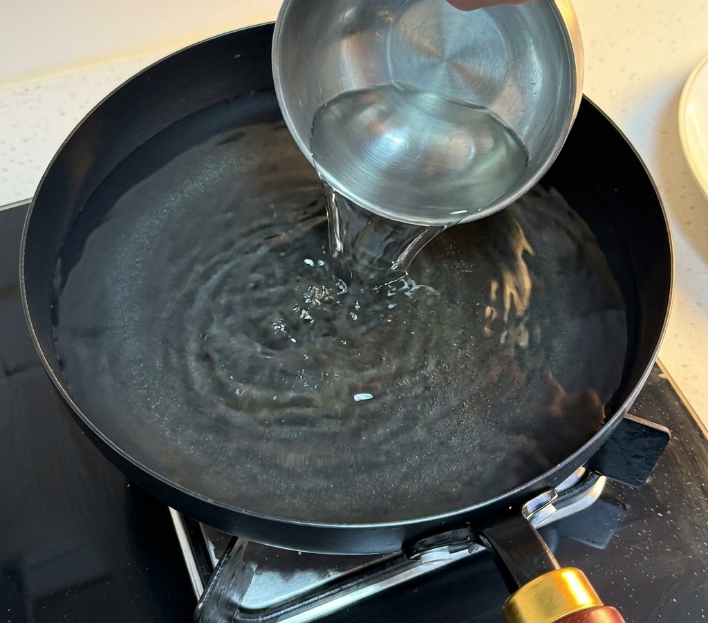 Eine Hand gießt klares Wasser aus einer Metallschüssel in eine Pfanne mit heißem Wasser.