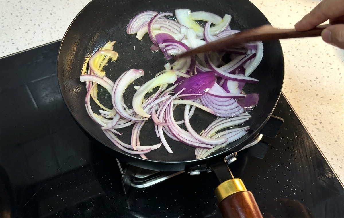 Rote Zwiebelstreifen werden mit einem Holzspatel in einer Pfanne angebraten.