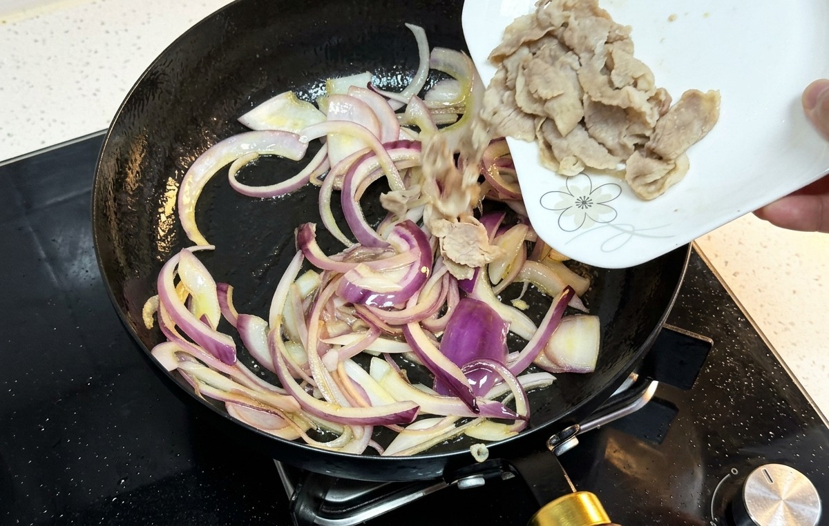 Gegartes Fleisch wird von einem Teller in die Pfanne zu den Zwiebeln gegeben.