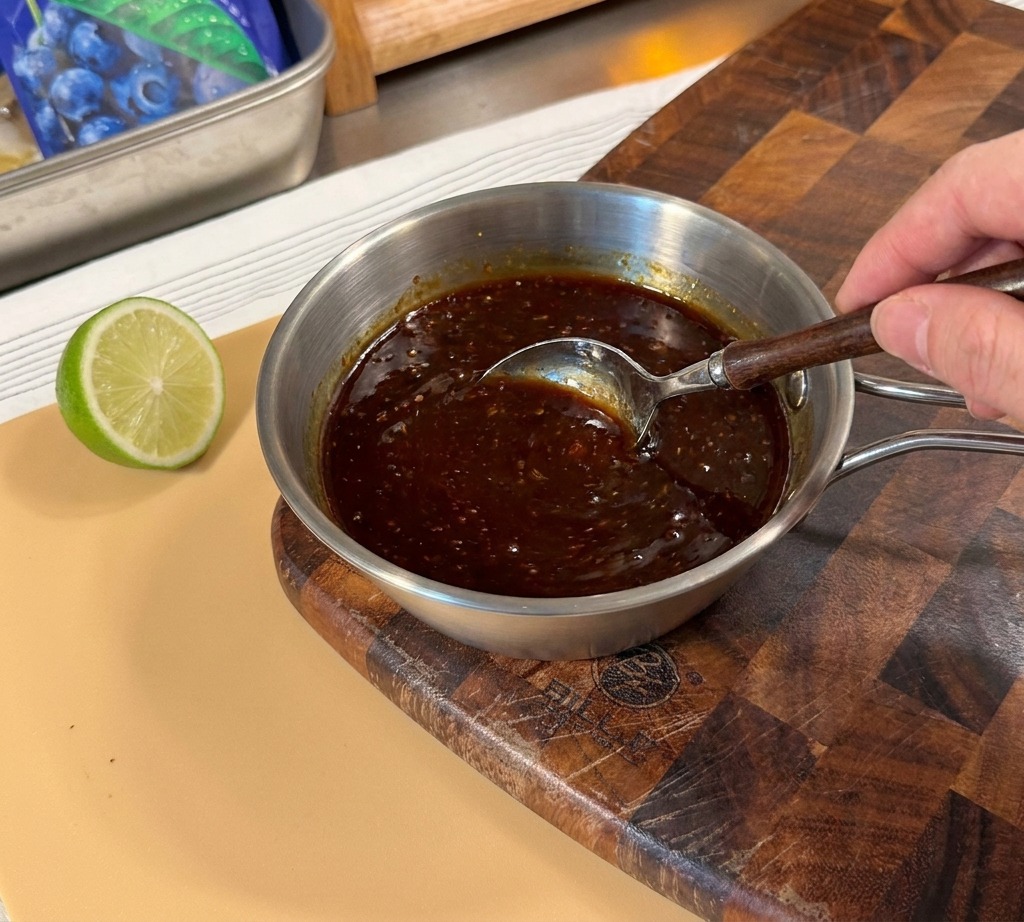 Eine Hand rührt mit einem Löffel eine dunkle Teriyaki-Saucenmischung in einem kleinen Metalltopf an.