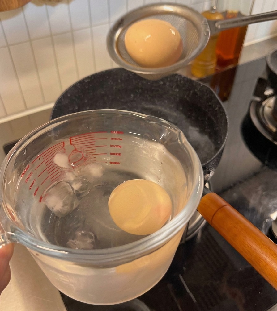 Ein gekochtes Ei wird aus dem Sieb in einen Messbecher mit Eiswürfeln und Wasser umgefüllt.