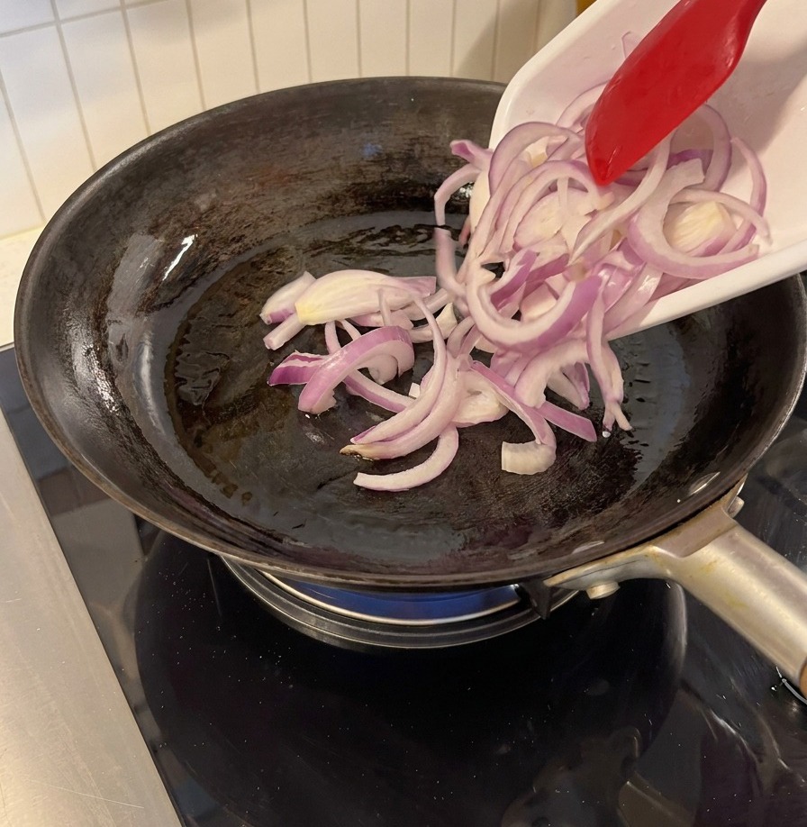 Rote Zwiebelstreifen werden mit einem roten Pfannenwender in einer schwarzen Pfanne angebraten.