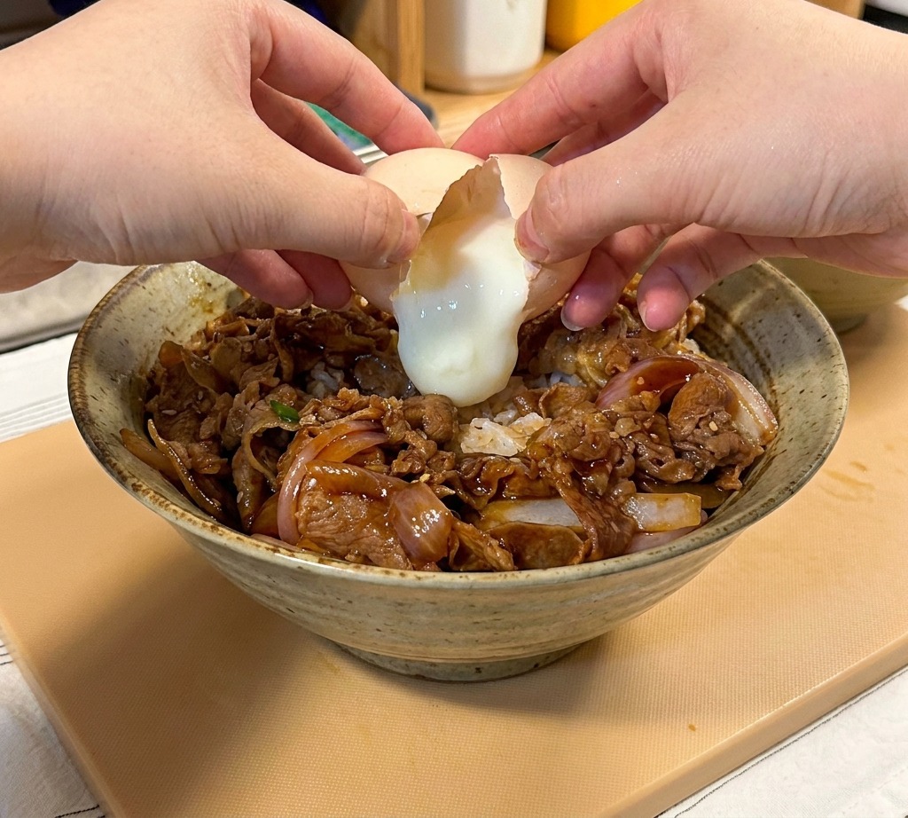Nahaufnahme von Händen, die ein weiches Onsen-Ei über der Gyudon-Schüssel aufschlagen.
