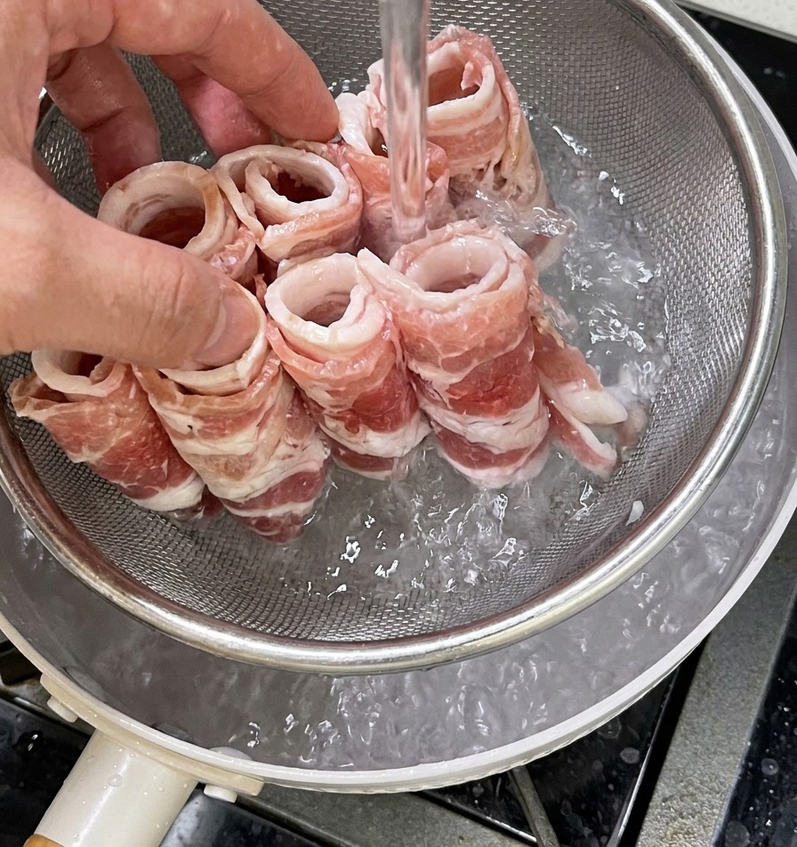 Eine Hand hält ein Metallsieb mit rohen Rindfleischrollen, das in einen Topf mit kochendem Wasser zum Blanchieren getaucht wird.