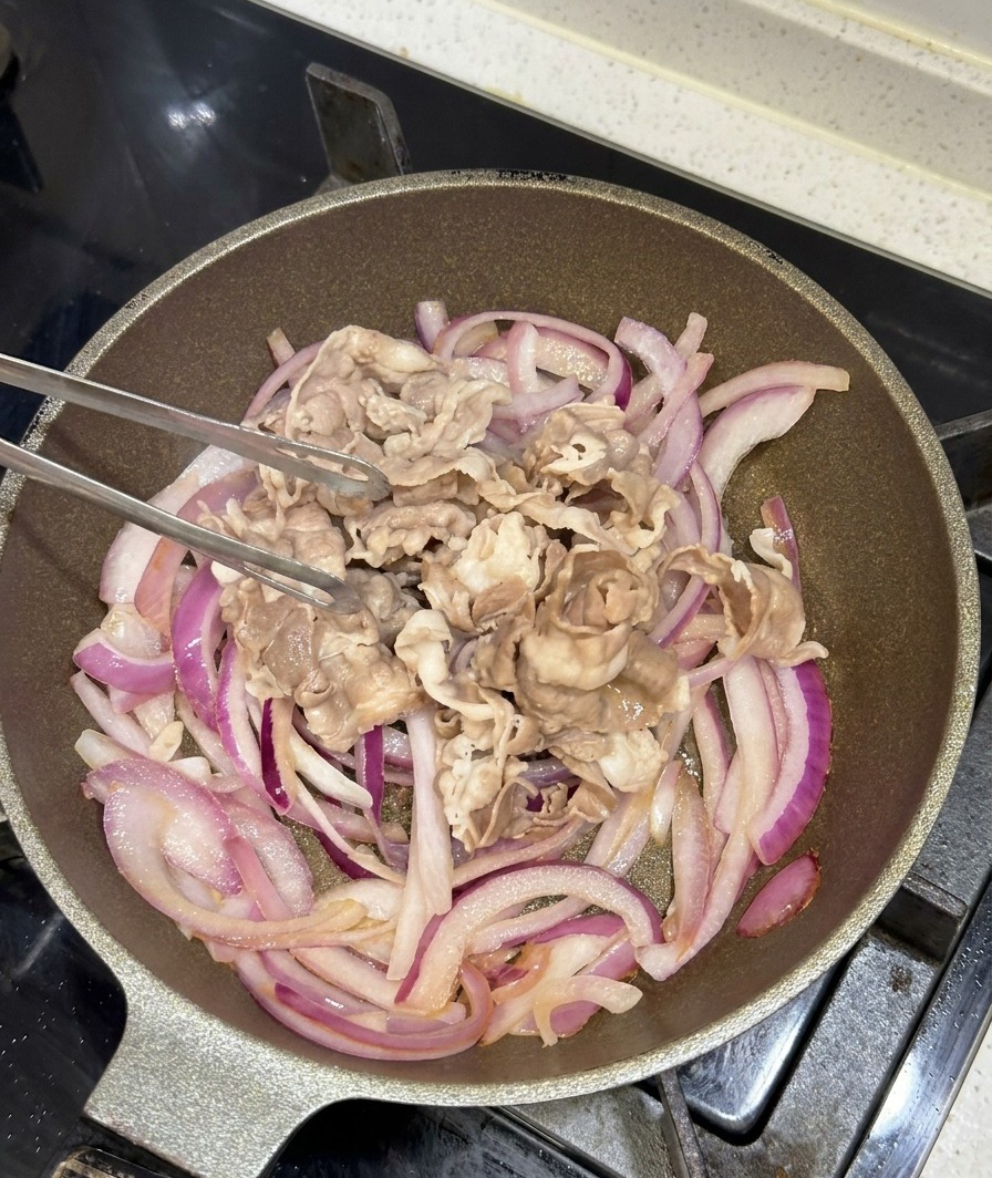 Blanchierte Rindfleischscheiben werden mit einer Metallzange in eine Pfanne mit angebratenen lila Zwiebeln gegeben.