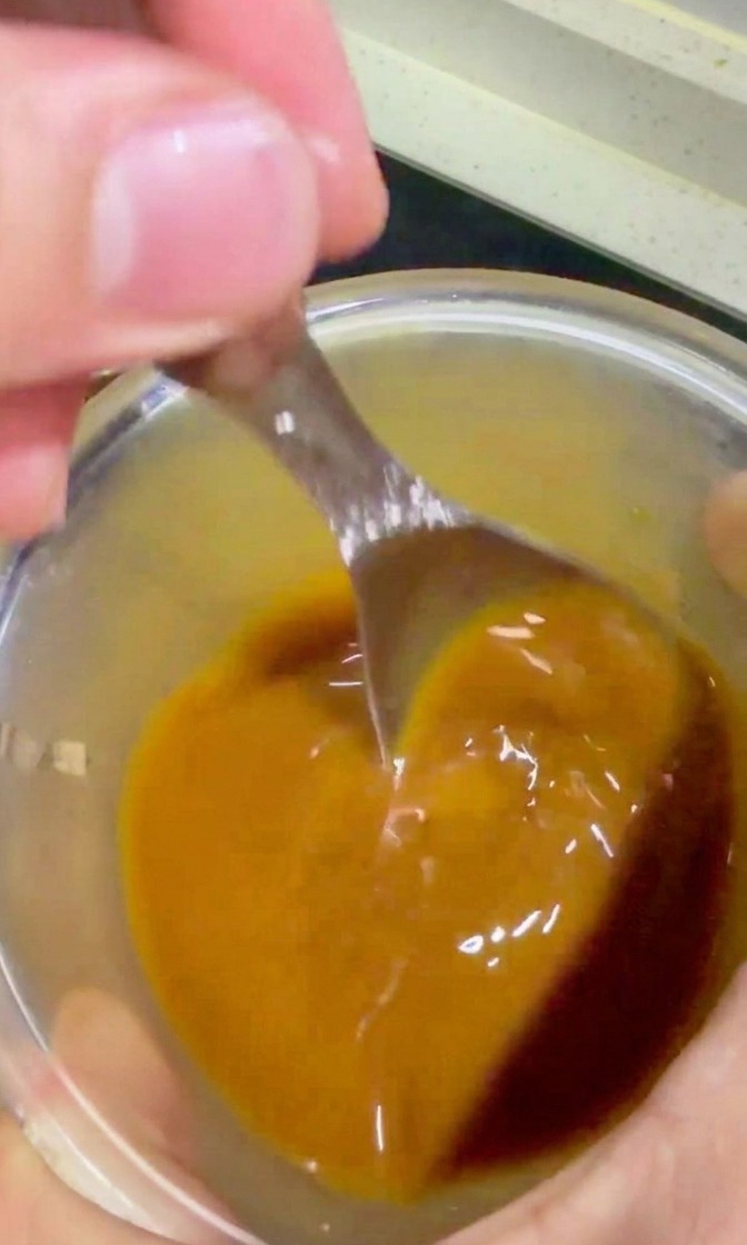 Eine Hand gießt mit einem Löffel eine goldbraune Teriyaki-Sauce aus einer Glasschale über das Fleisch und die Zwiebeln in der Pfanne.