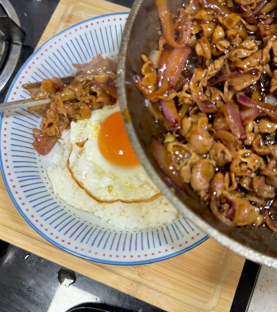 Glasiertes Teriyaki-Rindfleisch und Zwiebeln werden aus einer Pfanne in eine Schüssel mit Reis und Ei gegossen.