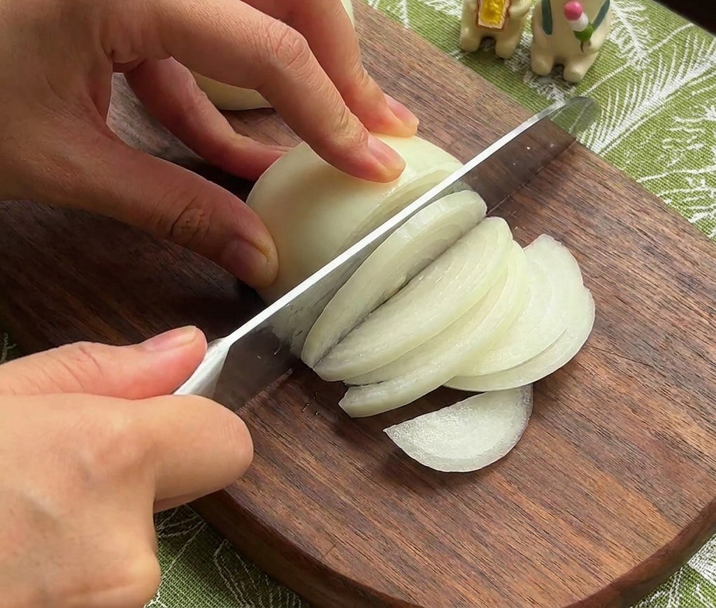 Hände schneiden eine weiße Zwiebel in feine Streifen auf einem dunklen Holzbrett.