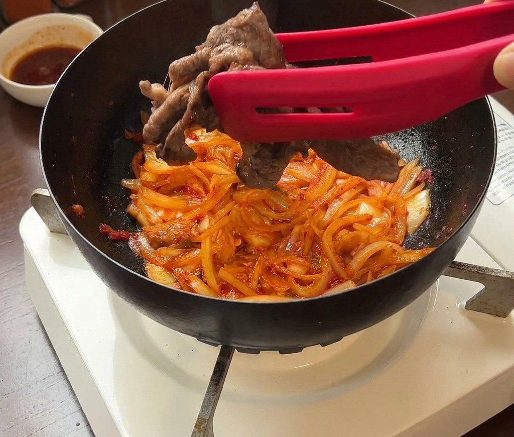 Rindfleischscheiben werden mit einer Zange zurück zum Kimchi in die Pfanne gegeben.