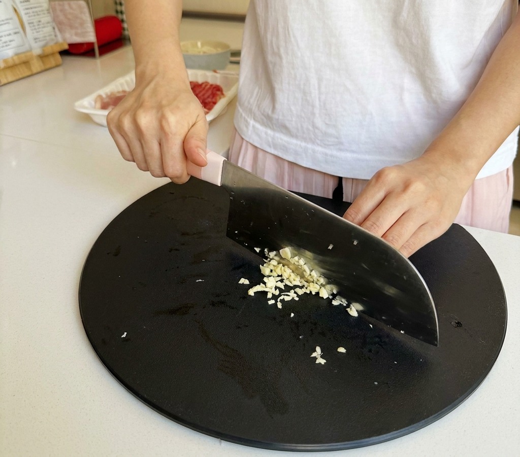Knoblauchzehen werden auf einem dunklen Schneidebrett mit einem Kochmesser fein gehackt.