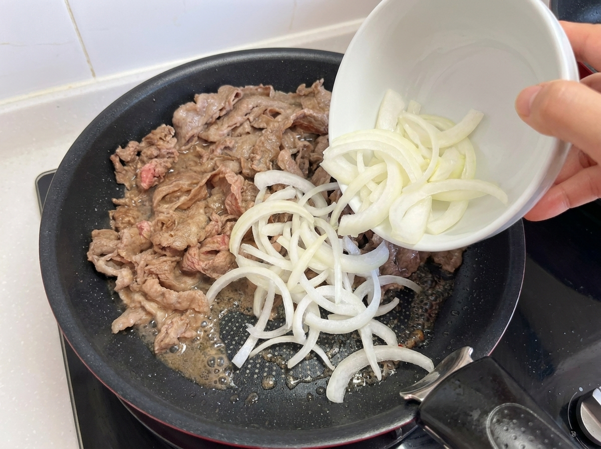 Zwiebelstreifen werden zum bereits angebratenen Fleisch in die Pfanne gegeben.