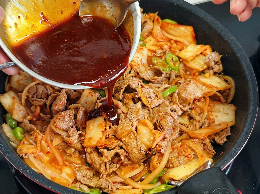 Die dunkelrote Gochujang-Sauce wird gleichmäßig über das Kimchi-Rindfleisch in der Pfanne gegossen.
