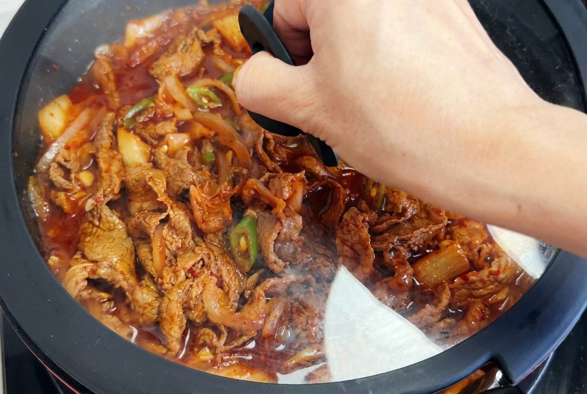 Ein Deckel wird auf die Pfanne gesetzt, in der das Fleisch in der roten Sauce schmort.