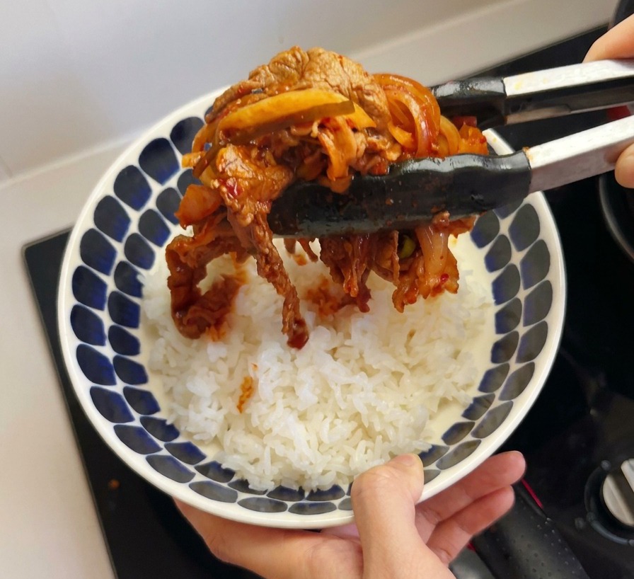 Das fertige Kimchi-Rindfleisch wird mit einer Küchenzange auf einer Schüssel mit weißem Reis angerichtet.