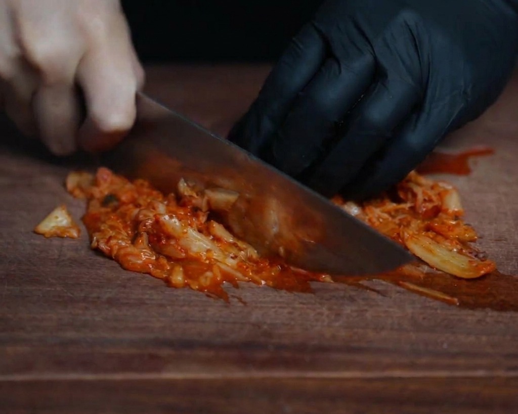 Nahaufnahme eines Kochs, der leuchtend rotes, fermentiertes Kimchi auf einem dunklen Holzbrett mit einem scharfen Messer fein hackt.