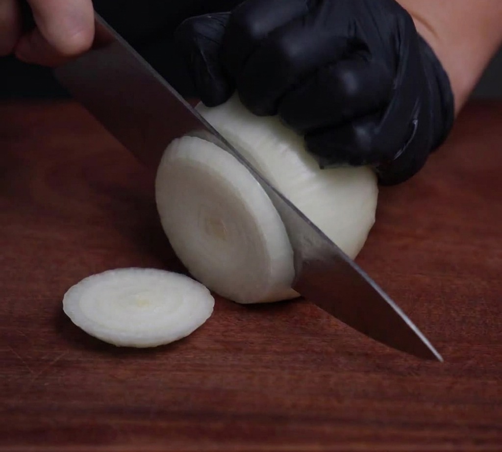 Eine weiße Zwiebel, die mit einem professionellen Kochmesser auf einer Holzoberfläche in dünne, durchscheinende Ringe geschnitten wird.