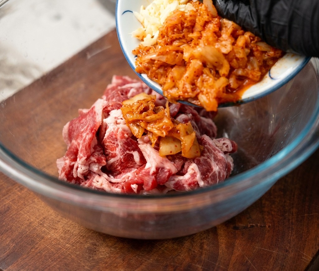 Gehacktes Kimchi und fein gehackter Knoblauch werden von einem kleinen Keramikteller in eine Schüssel mit rohem, dünn geschnittenem Rindfleisch gegeben.