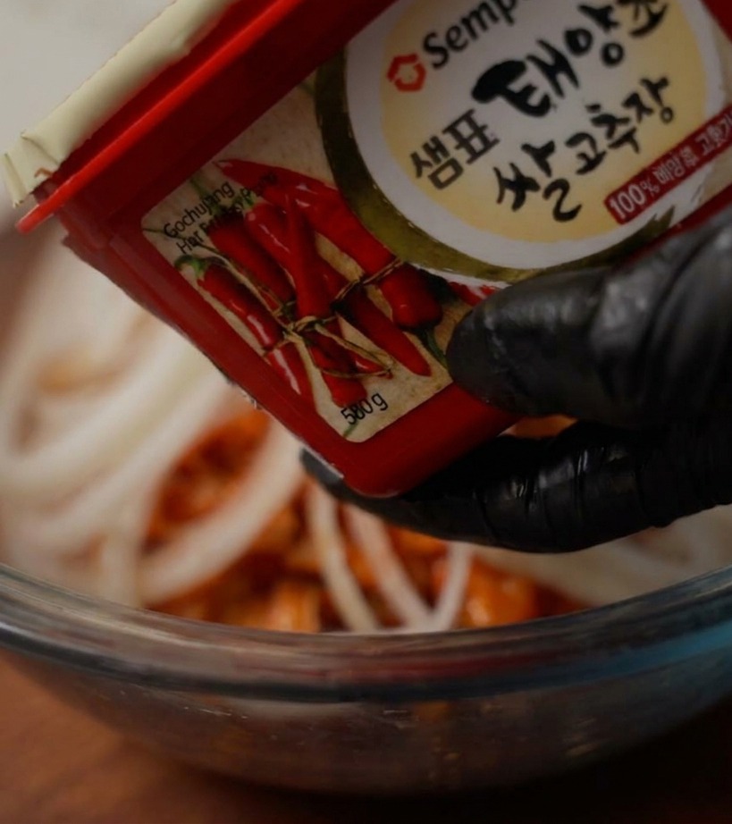 Eine Hand in einem schwarzen Handschuh hält einen Behälter mit roter Chilipaste über eine Schüssel, um eine Portion Paste hinzuzufügen.