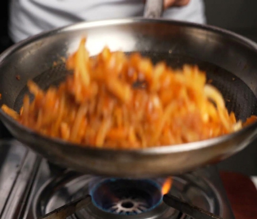 Rindfleisch und Zwiebeln werden in einer Metallpfanne mit einem Holzspatel über einer Gasflamme geschwenkt und gebraten.