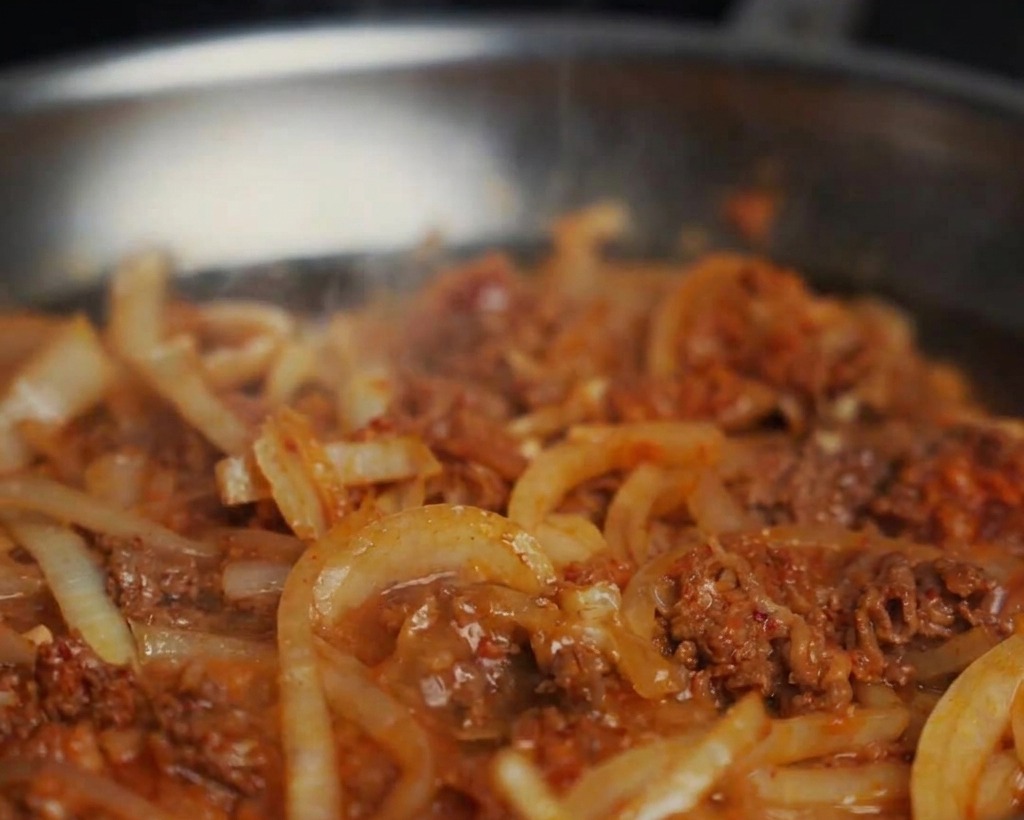 Wasser wird in eine Pfanne mit gebratenem Rindfleisch und Zwiebeln gegossen, wodurch eine blubbernde Sauce entsteht.
