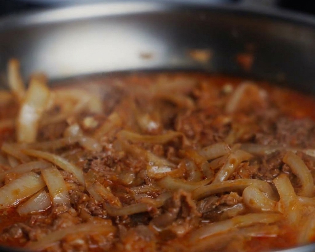 Nahaufnahme von schwarzem Pfeffer und Sojasauce, die in eine köchelnde Pfanne mit Rindfleisch und Zwiebeln gegeben werden.