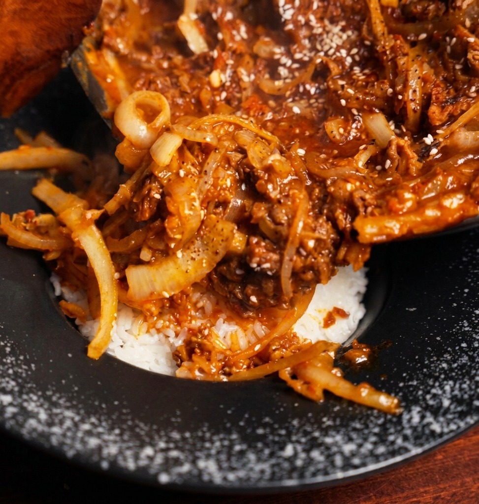 Herzhaftes Rindfleisch-Zwiebel-Ragout wird aus einer Pfanne auf ein Bett aus lockerem weißem Reis in einer schwarzen Schüssel gegeben.
