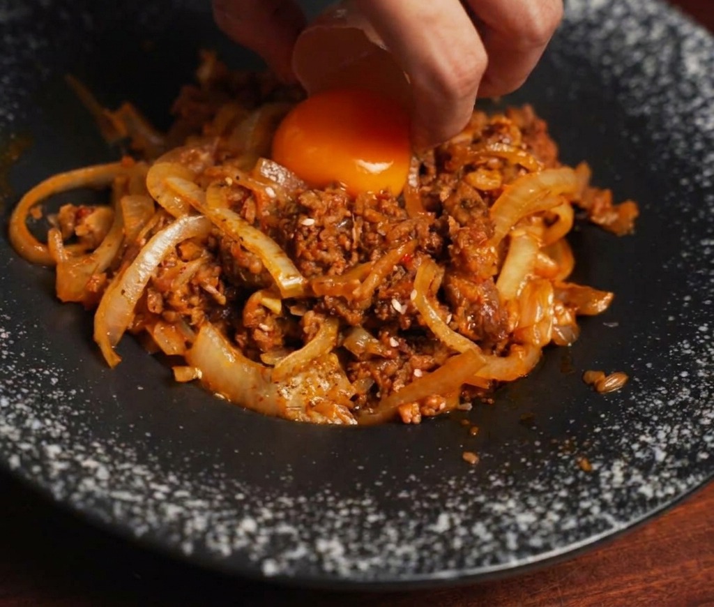 Eine Hand lässt mithilfe einer Eierschale vorsichtig ein leuchtend orangefarbenes Eigelb auf einen Berg aus gebratenem Rindfleisch und Zwiebeln gleiten.