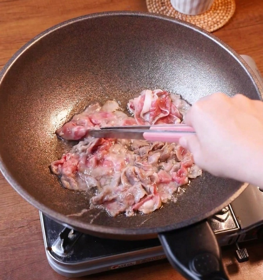 Gebräunte Rindfleischscheiben werden mit einer Grillzange aus einer dunklen Pfanne gehoben.