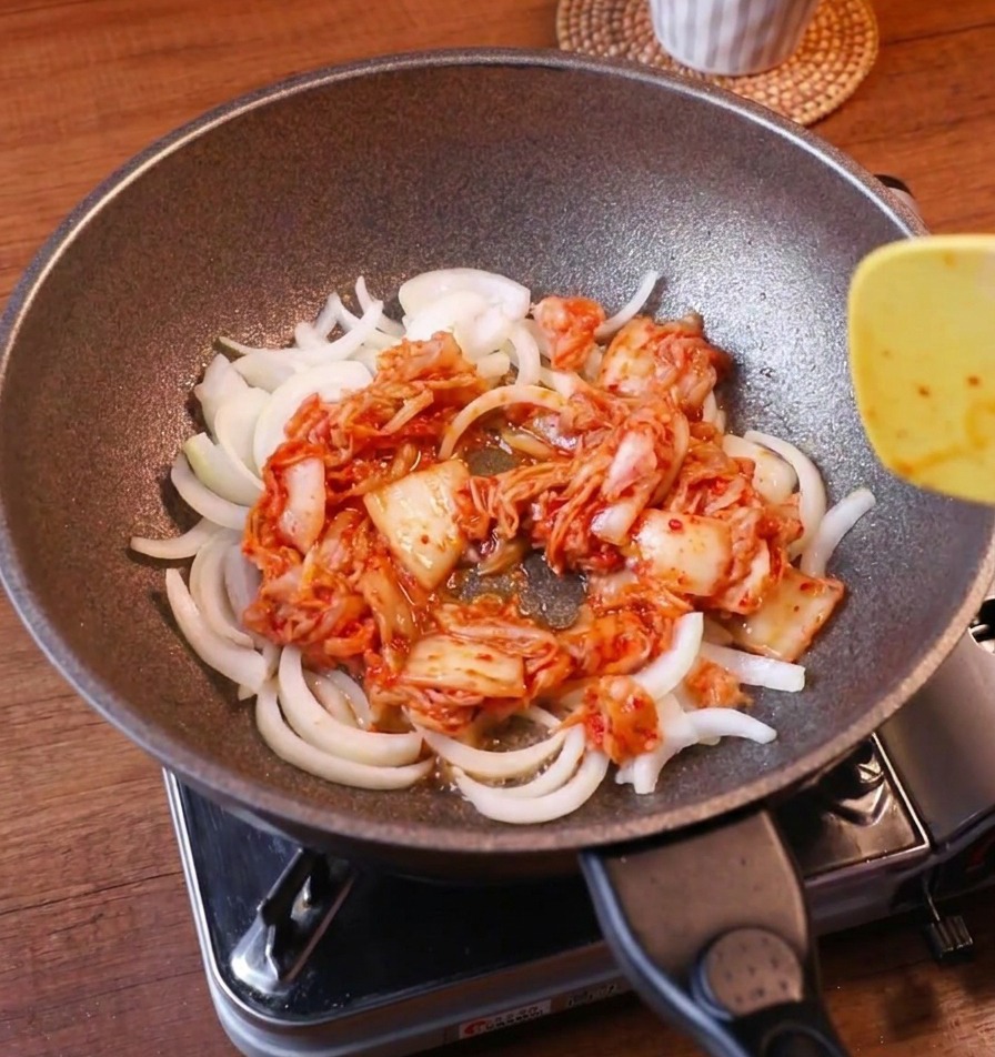 Weiße Zwiebeln und leuchtend rotes Kimchi werden mit einem gelben Pfannenwender zusammen angebraten.