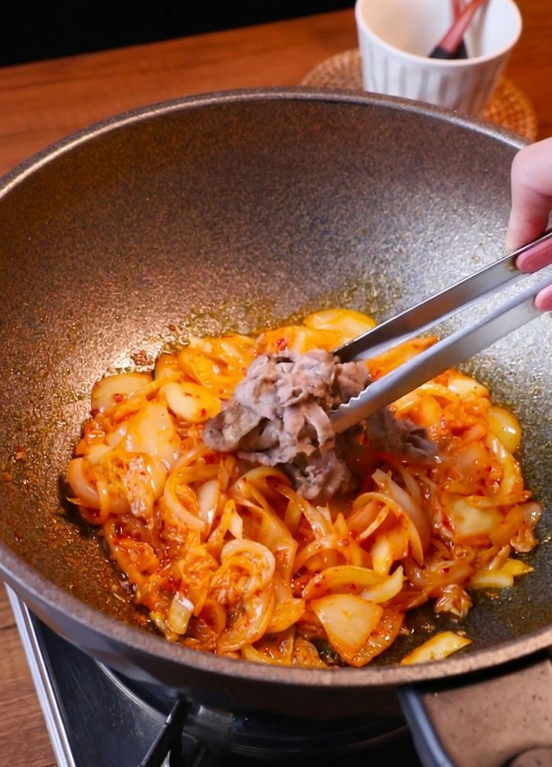 Eine Zange legt die gebräunten Fleischstreifen zurück in die Pfanne zu Kimchi und Zwiebeln.