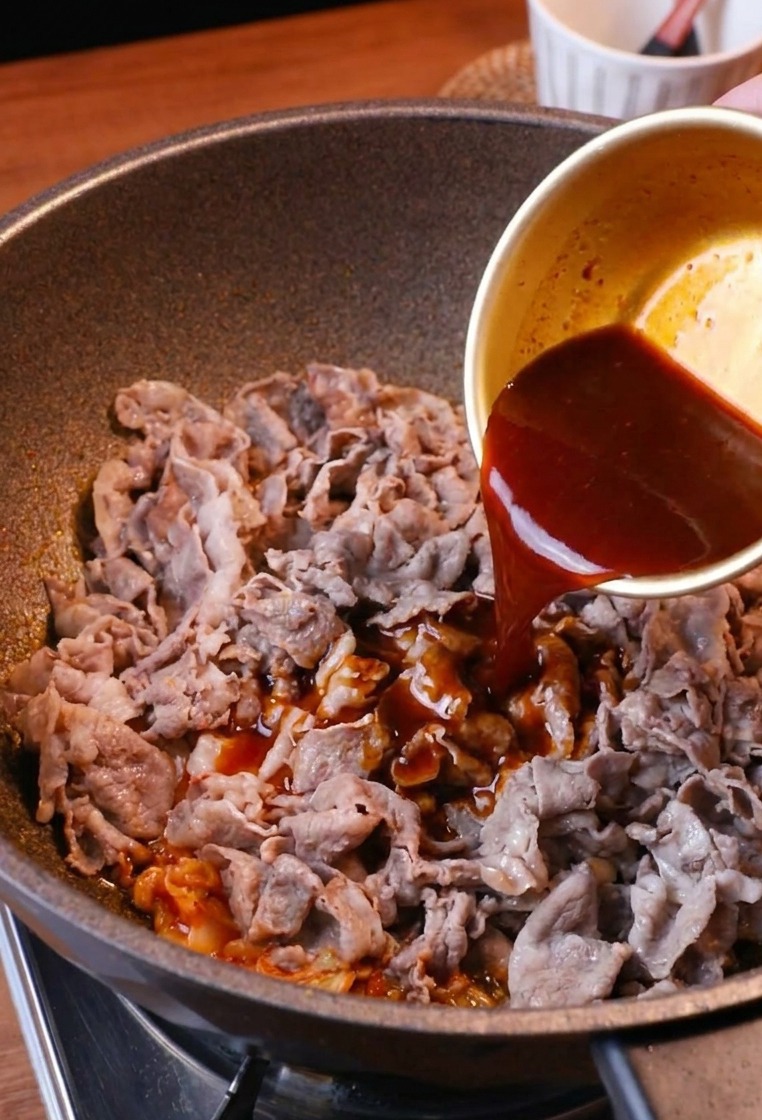 Eine rötlich-braune Sauce wird aus einer Schüssel über das Fleisch und den Kimchi in der Pfanne gegossen.