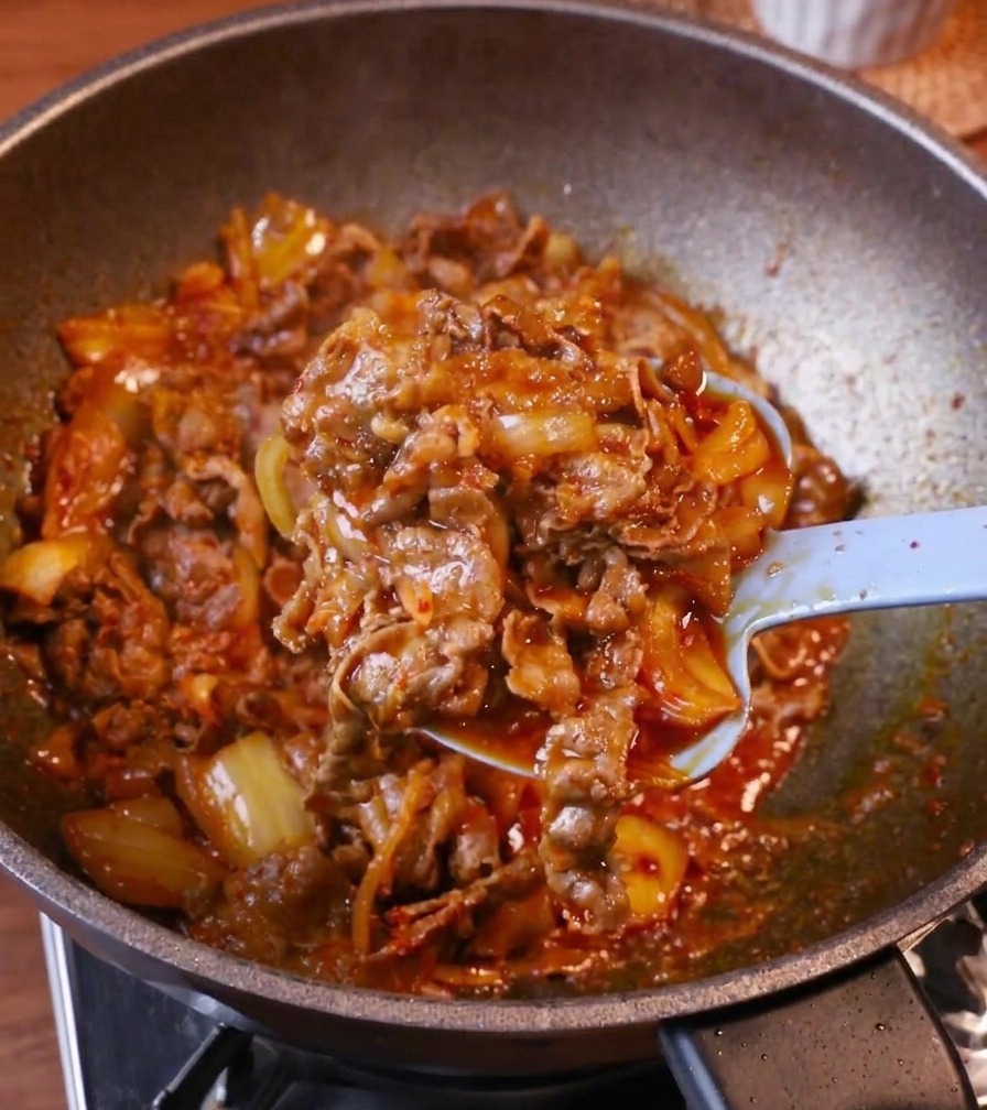 Ein hellblauer Löffel schichtet eine großzügige Portion Kimchi-Fleisch auf eine Schüssel mit dampfendem Reis.