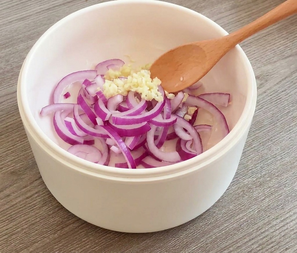 Rote Zwiebelringe und gehackter Knoblauch werden mit einem Holzlöffel in einer weißen Pfanne gerührt.