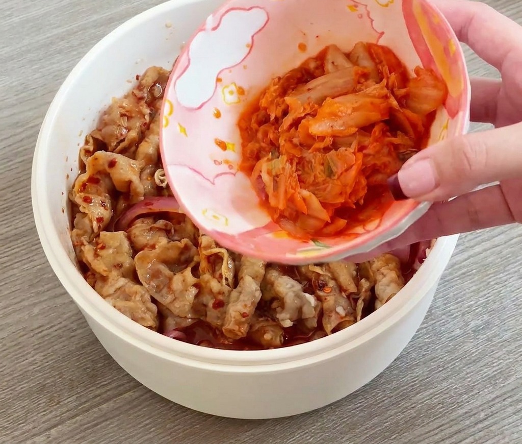 Eine Hand gibt eine Schale mit geschnittenem Kimchi in die Pfanne zum Fleisch.