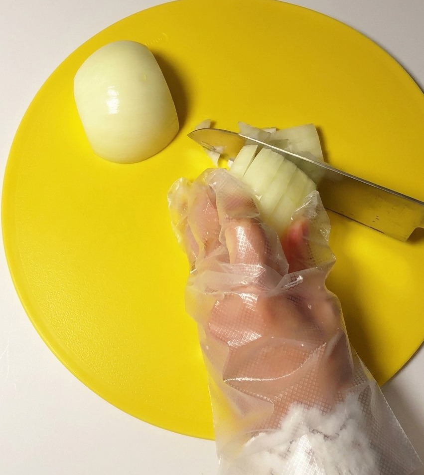 Eine Hand mit Plastikhandschuh würfelt eine weiße Zwiebel mit einem Edelstahlmesser auf einem gelben Schneidebrett.