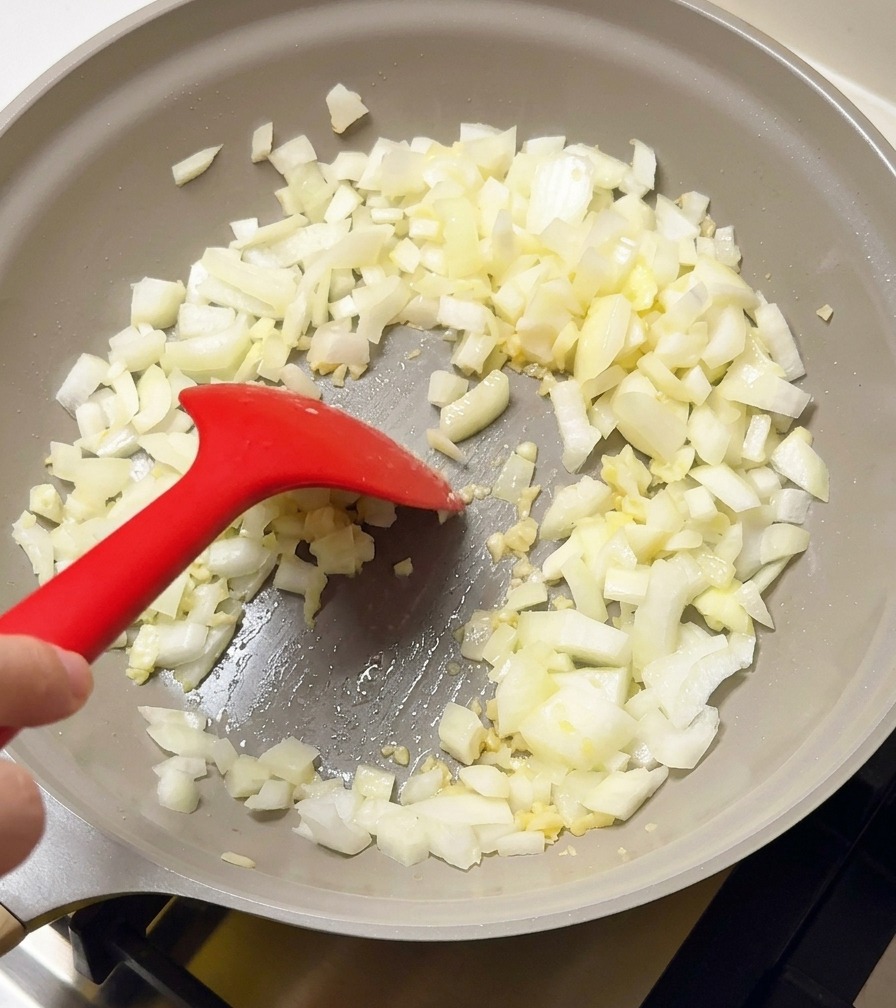 Gewürfelte Zwiebeln und Knoblauch werden in einer hellgrauen Pfanne mit einem roten Silikonspatel angebraten.